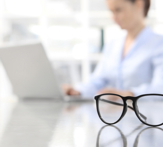 Closeup einer Brille am Schreibtisch, Frau im Hintergrund